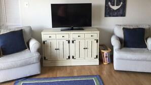 Ein Wohnzimmer mit Fernseher auf einer Kommode, zwei gestreiften Sesseln, einem blauen Teppich und einem Wandbild mit Anker-Motiv.