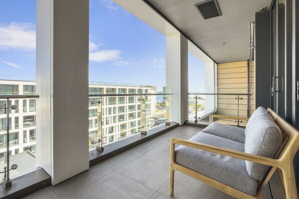 A balcony with a view of modern buildings, a grey sofa, and a wooden-framed chair.