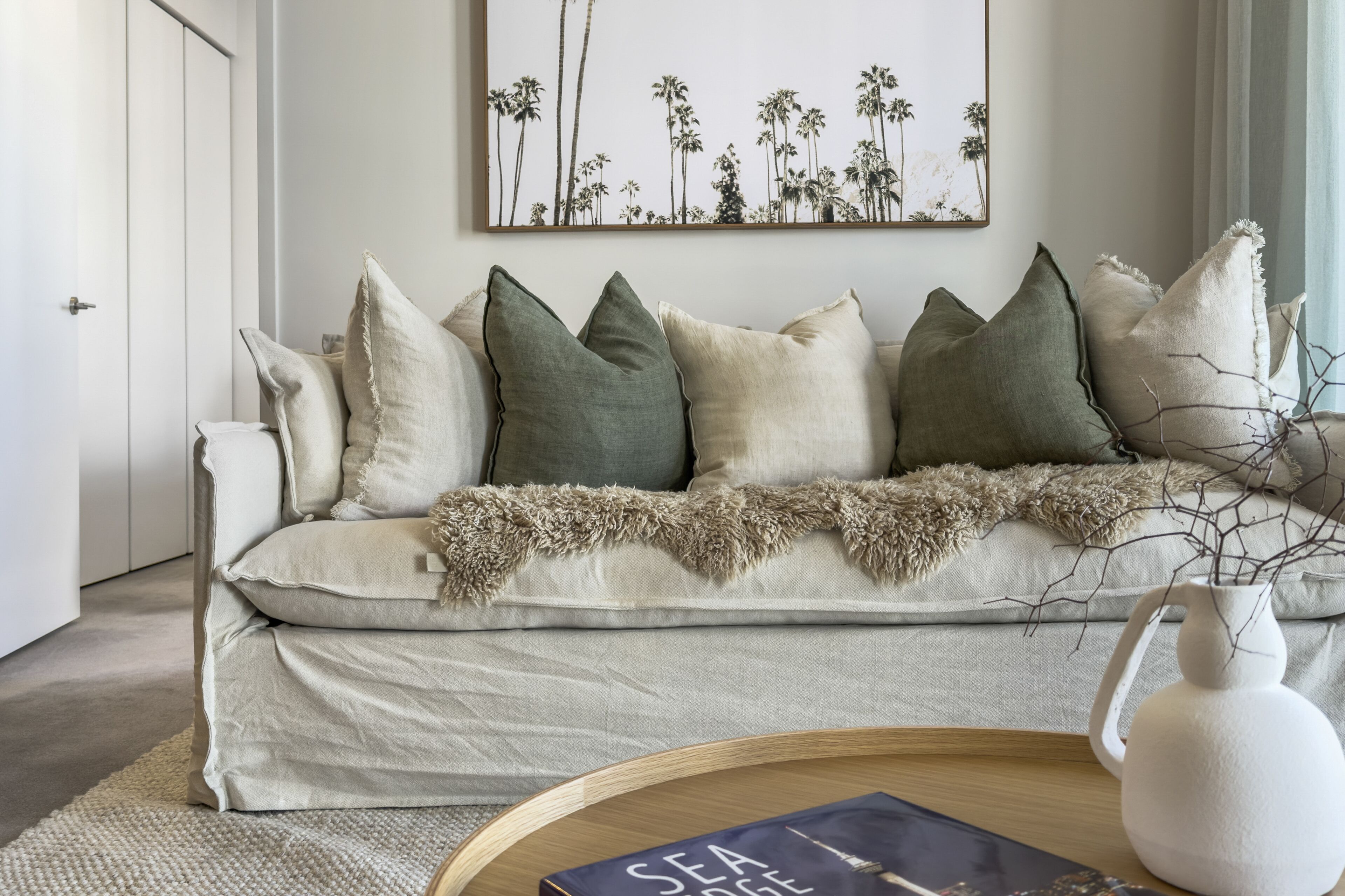 Un salon avec un canapé beige, plusieurs coussins, un plaid, un vase blanc et un tableau encadré accroché au mur.