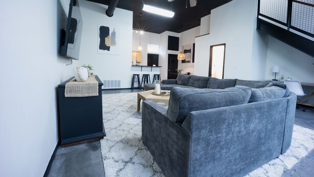 Una sala de estar moderna con un sofá gris, una mesita y un ventilador de techo. Se ve una escalera que sube a un nivel superior.