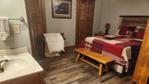 A bedroom with a wooden floor, a bed with a red and black checkered blanket, a wooden bench, a nightstand with a lamp, and a painting on the wall.