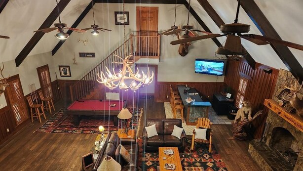 A spacious loft-style room with a high ceiling, wooden beams, a fireplace, a pool table, leather seating, and a television.