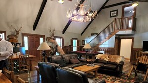 A spacious living room with leather sofas, a wooden coffee table, and a patterned rug. A chandelier hangs from the ceiling, and a staircase is visible in the background.