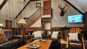 A spacious living room with a high ceiling, a wooden staircase, a mounted deer head, and a television on the wall.