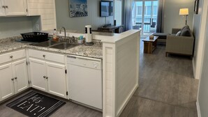A modern kitchen with white cabinets, a granite countertop, and a built-in dishwasher. There is a dining area with a sofa and a lamp.
