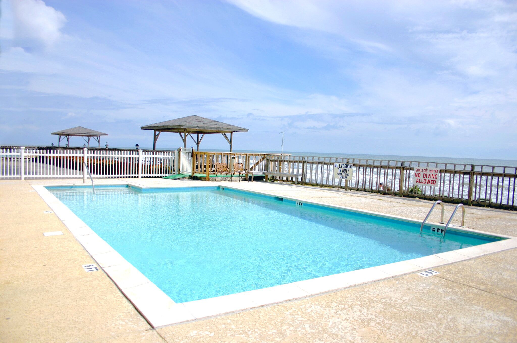 A rectangular swimming pool with a white deck, two shaded areas, and a white fence along the edge.