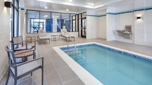 A modern indoor pool area with a clear view through large windows, a pool with a blue tiled edge, and a seating area with chairs and a table. - Hyatt Place Newark - Main Street (Newark)