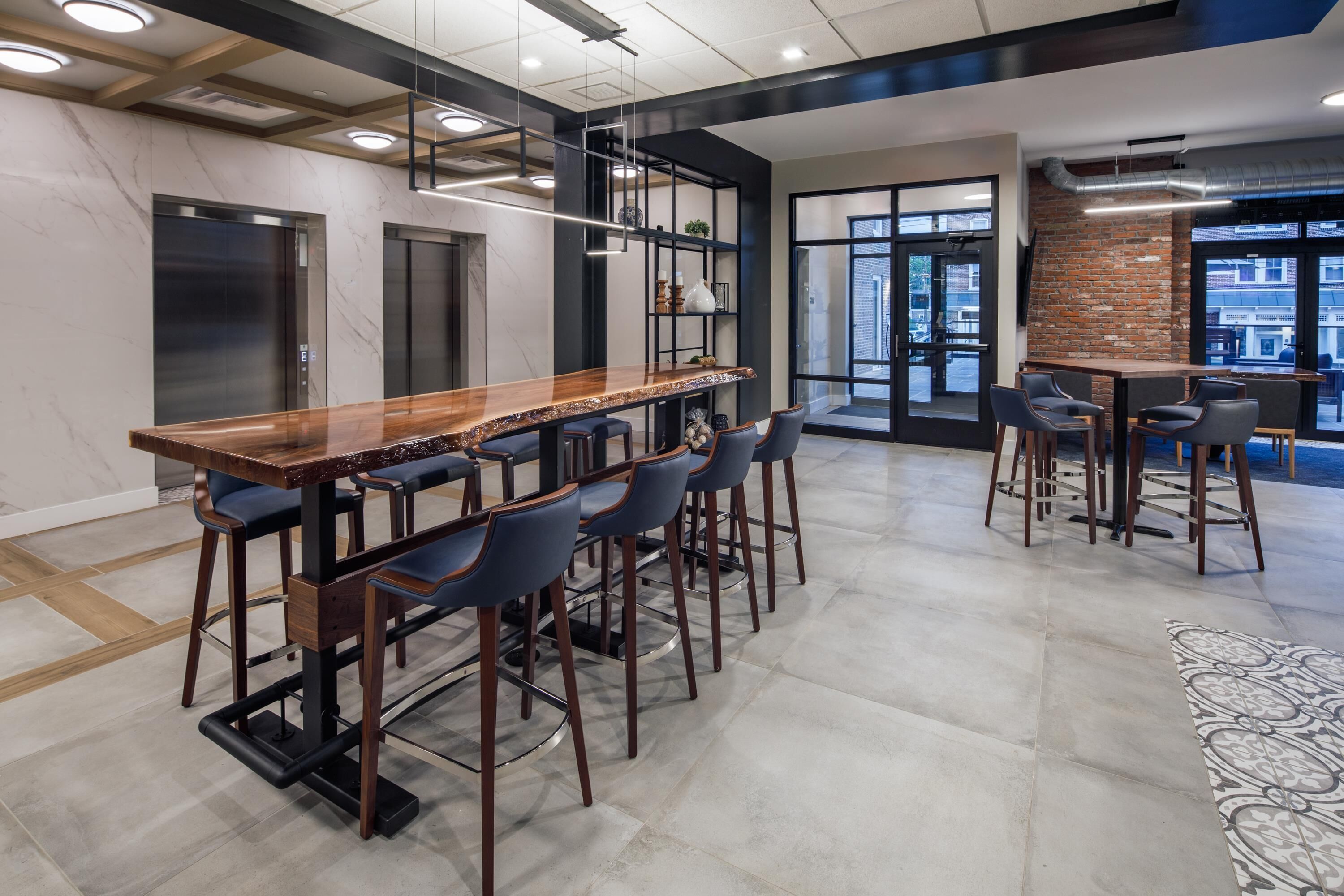 A modern bar area with a long wooden counter, multiple bar stools, and a view of the city through large windows.