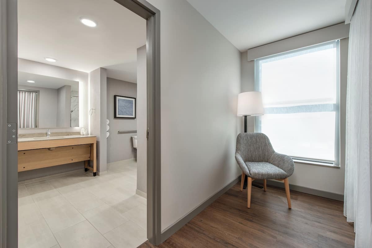 Una habitación de hotel moderna con suelo de madera, un ventanal con cortinas, un sillón gris y un baño con tocador de madera y un espejo grande.
