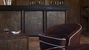 A bar area with a dark wooden bar, shelves of bottles, a cushioned chair, and a small table with a glass and a bottle. - Villa Al Assala La Palmerale (Marrakech)