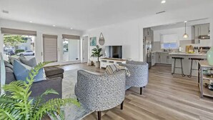 A modern living room with a sofa, armchairs, and a TV. There is a kitchen area with a dining table and bar stools.