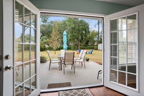 Un patio abierto con una mesa y sillas, una sombrilla azul y vista a un jardín con árboles y césped.