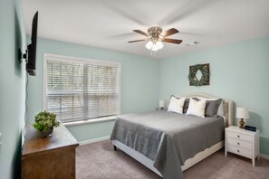 Un dormitorio con cama, mesitas de noche, ventilador de techo, ventana con persianas, televisor de pared y una planta pequeña.