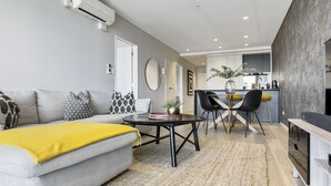 A modern living room with a grey sofa, yellow throw, and a dining area with a round table and chairs. - Laid back living with city views (Auckland)