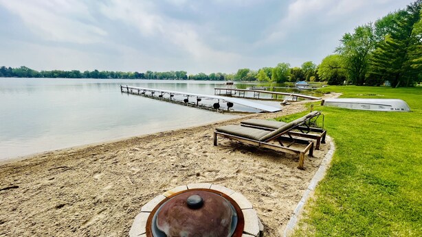 En strand vid sjön med solstolar, en grill och en brygga som sträcker sig ut i vattnet.