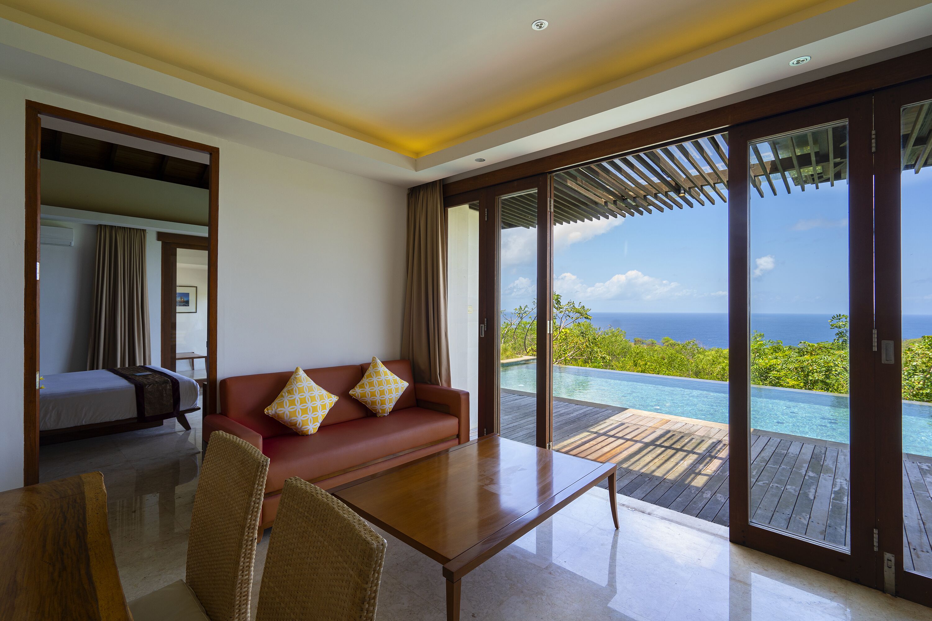 A modern hotel room with a wooden floor, a red sofa with patterned cushions, a wooden coffee table, and a large sliding glass door leading to a private pool with a view of the ocean.