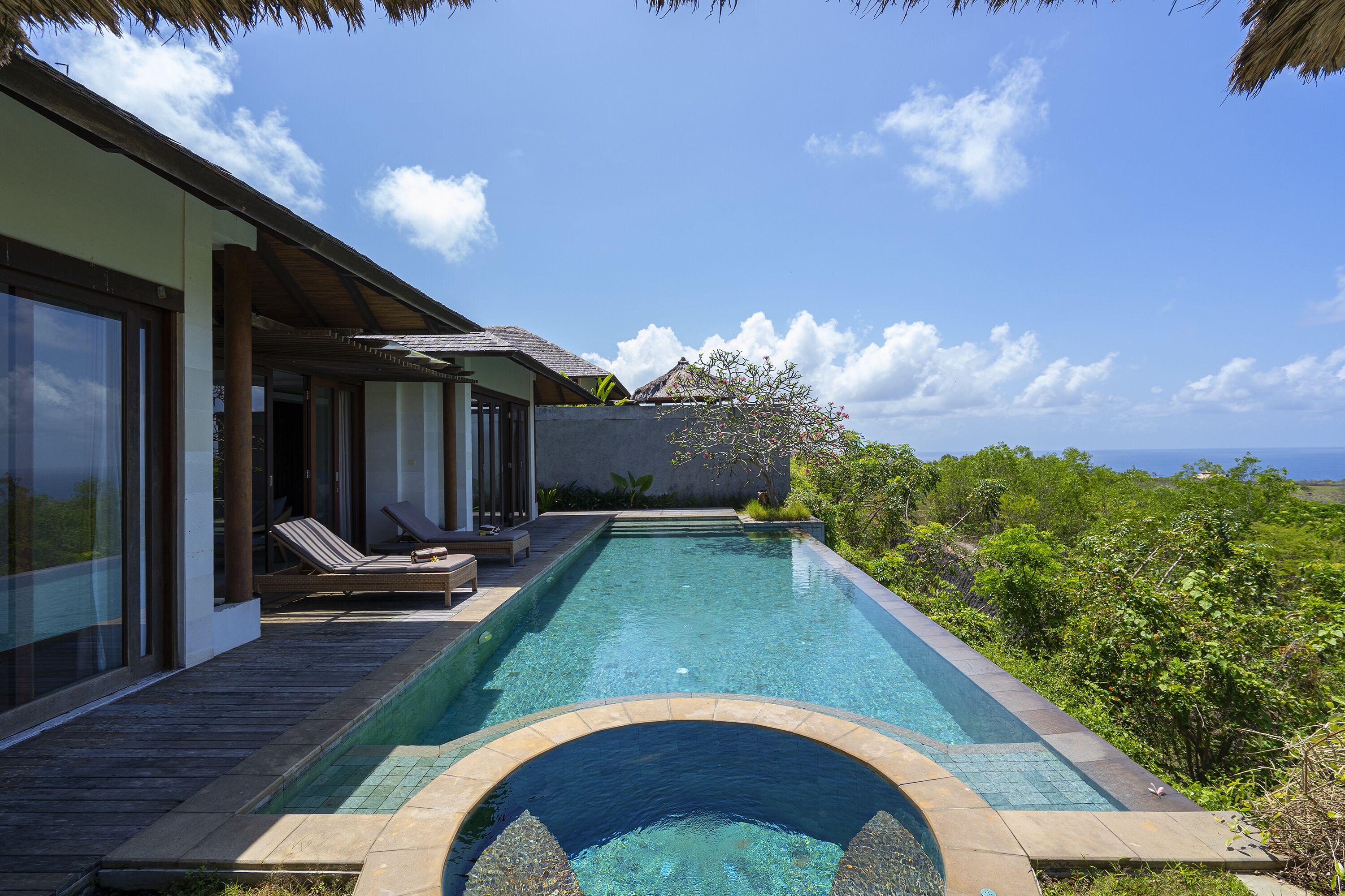 A swimming pool with a clear view of the ocean, a covered patio with lounge chairs, and a lush green landscape.
