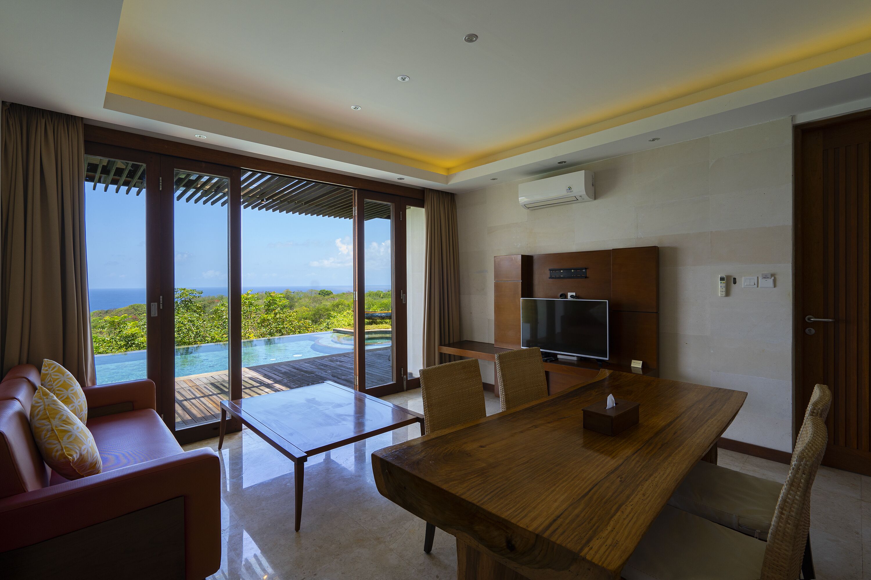 A modern hotel room with a wooden dining table, comfortable seating, a flat-screen TV, and a large sliding glass door leading to a pool with a scenic view.