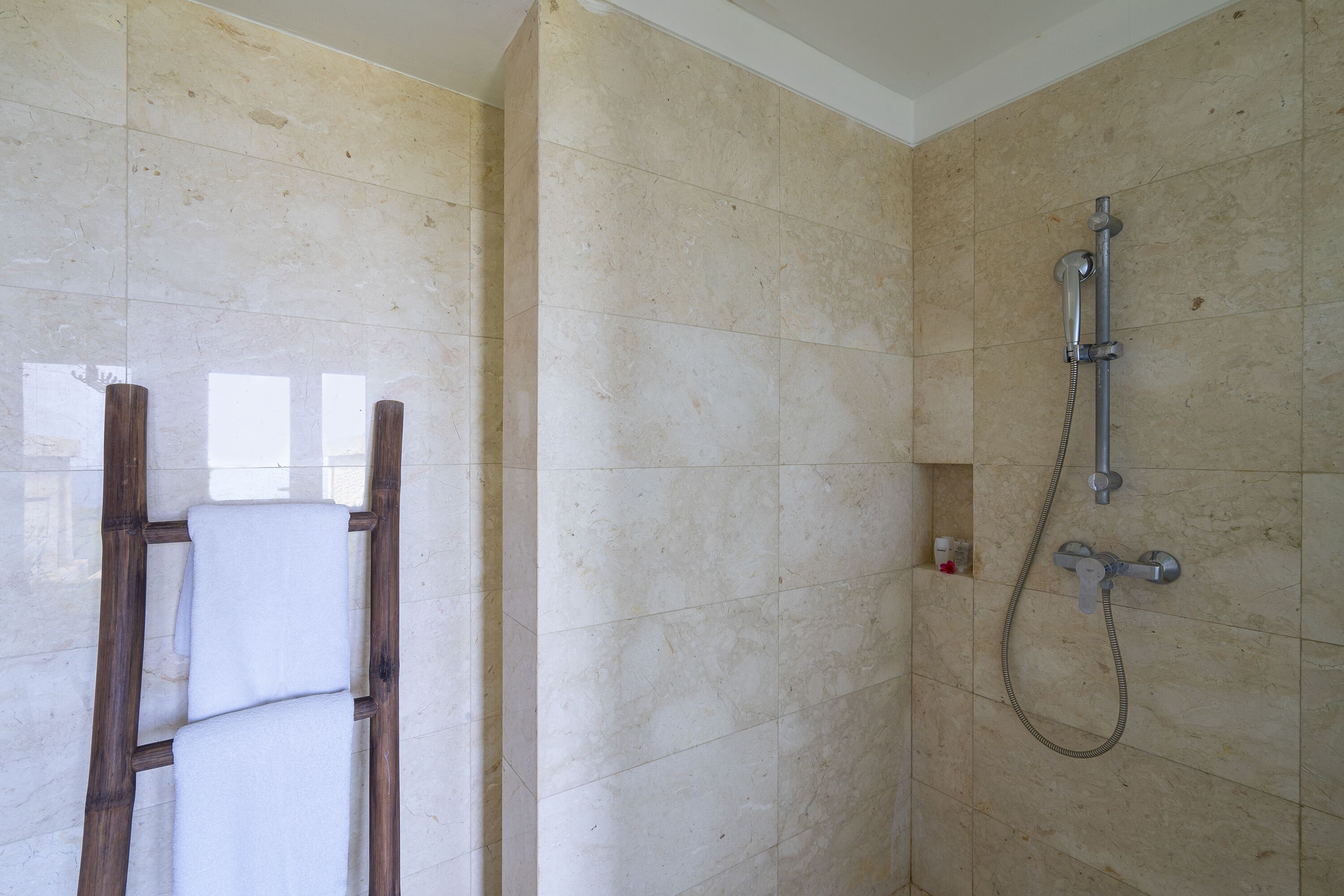 A bathroom with beige tiled walls, a shower with a handheld showerhead, and a wooden ladder leaning against the wall.