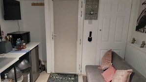 Ein kompakter Wohnbereich mit Küchenzeile, einem grauen Sofa mit Zierkissen, einem an der Wand montierten Fernseher und einer Tür mit dem Schriftzug "TAKE LIFE ONE CUP AT A TIME".