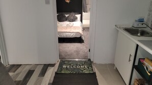 Ein Flur, der in ein Schlafzimmer mit einem Bett, einer kleinen Küchenzeile und einer Fußmatte führt.