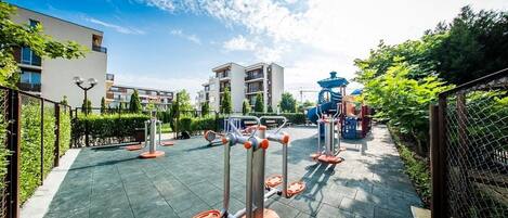An outdoor exercise area with fitness equipment, surrounded by greenery and buildings.