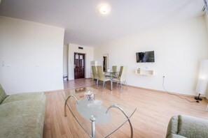A modern living room with a glass coffee table, a flat-screen TV mounted on the wall, and a dining area with chairs and a table. - Orchid Fort Garden (Nessebar)