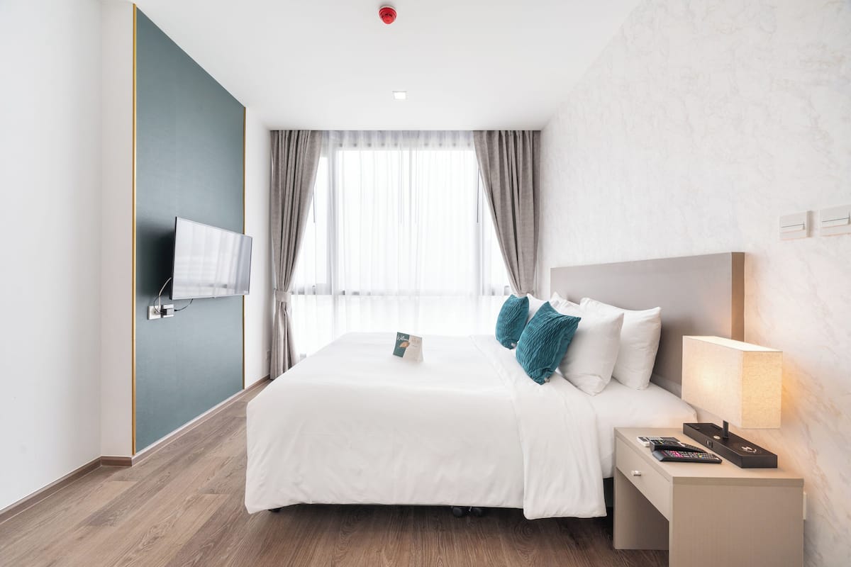Ein Hotelzimmer mit einem großen Bett, einem Nachttisch mit Lampe, einem an der Wand montierten Fernseher und einem Fenster mit Vorhängen.