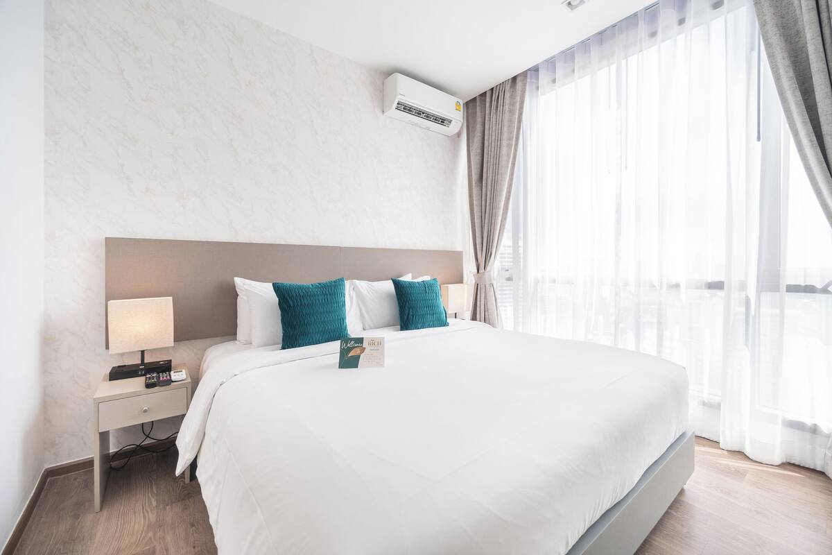 Ein Hotelzimmer mit einem großen Bett, einem Nachttisch mit Lampe, einer Klimaanlage und durchsichtigen Vorhängen.