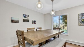 A dining room with a wooden table and benches, framed artwork on the walls, and a large window with a view of greenery. - Hudson Valley Hilltop Escape w/Hot Tub, 5private acres, near wine trail, orchard (Marlboro)