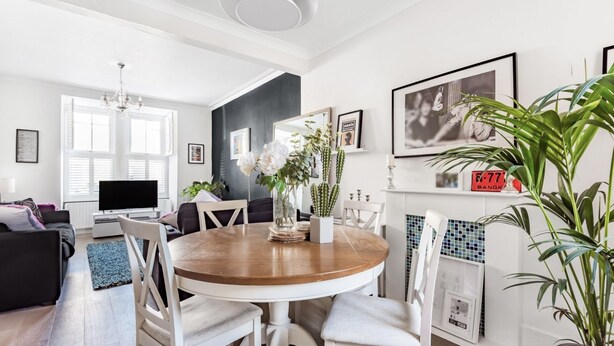 A modern living room with a round wooden dining table, white chairs, a black sofa, and a television. - Inviting 3-bed House in Beckenham (Beckenham)