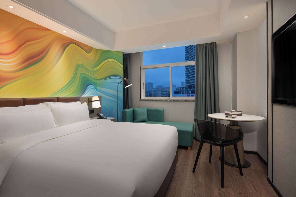 A hotel room with a large bed, a green sofa, a small table, and a view of a cityscape through a window.