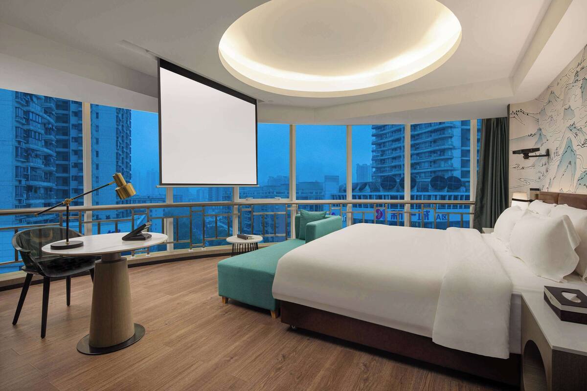 A hotel room with a large bed, a desk with a lamp, a small table, and a view of tall buildings through large windows.