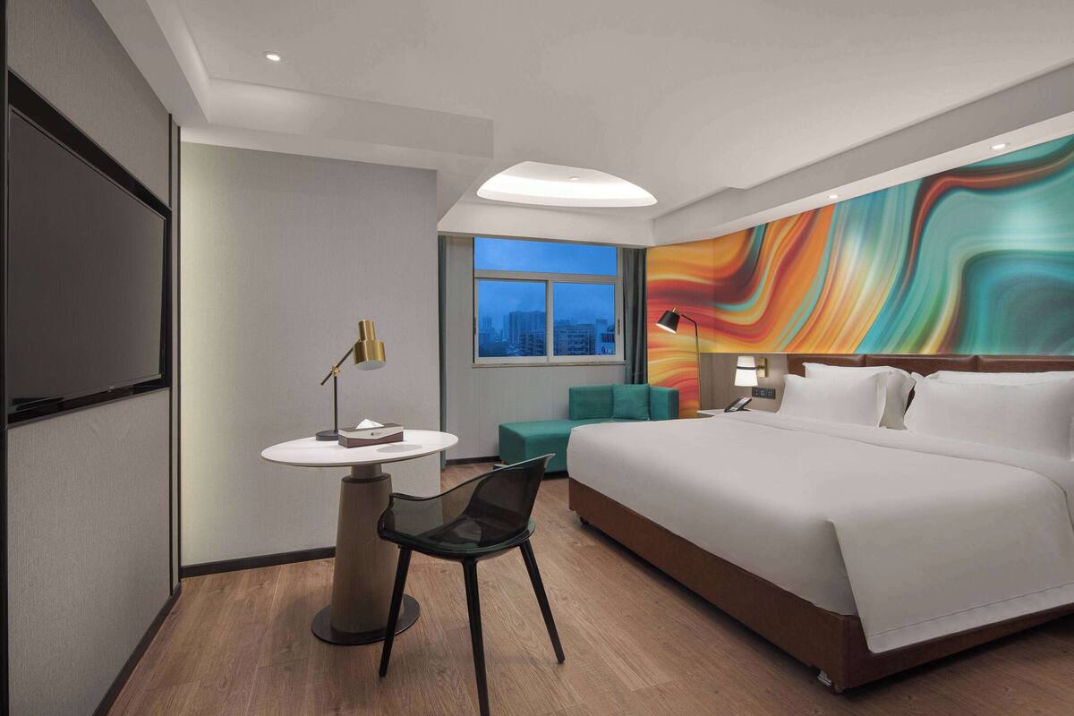 A modern hotel room with a large bed, a desk with a chair, a round table, and a colorful abstract wall mural.