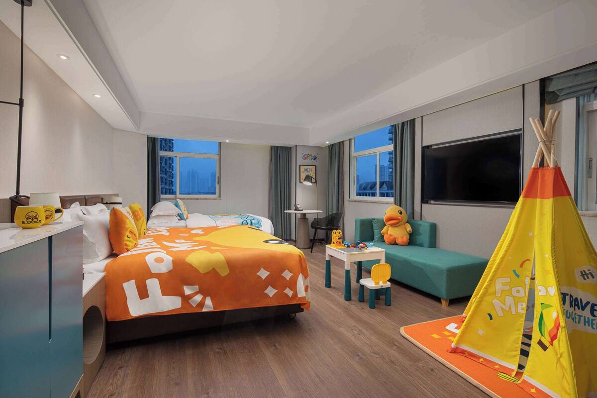 A modern hotel room with a large bed, a small table, a chair, a sofa, and a play tent.