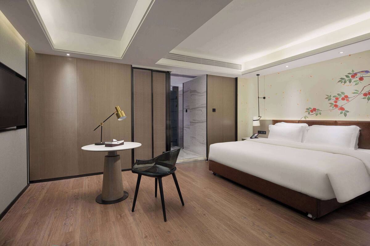 A hotel room with a bed, a desk with a chair, a small table, and a wall with a floral mural.