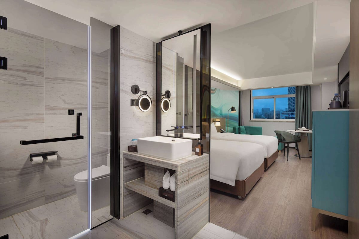 A modern hotel room with a glass-enclosed shower, a large bed, a desk, and a view of the cityscape.