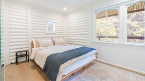 Een slaapkamer met een bed, nachtkastje, plafondventilator, raam met uitzicht op bomen en een wand met horizontale lambrisering.