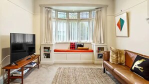 A living room with a large flat-screen TV, a sofa, a bench with cushions, a rug, and a window with curtains.