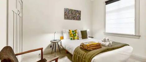 Un dormitorio con cama, una silla, una mesita de noche, una lámpara, una ventana y un cuadro en la pared.