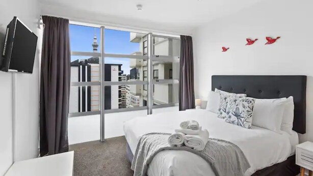 A modern hotel room with a large bed, a flat-screen TV, and a city view through floor-to-ceiling windows. - Central City 1Br Apartment (Auckland)