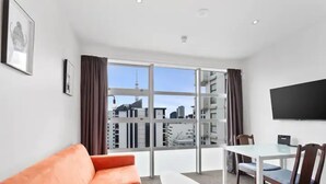 A modern hotel room with a large window, a sofa, a dining table with chairs, and a flat-screen TV mounted on the wall. - Central City 1Br Apartment (Auckland)