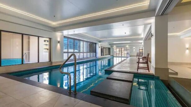 Une grande piscine intérieure à l’eau d’un bleu limpide, entourée de murs recouverts de carreaux et bordée d’une longue zone avec des marches menant à l’eau.