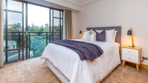 Une chambre à coucher avec un grand lit, une table de chevet, une lampe et une vue sur la ville grâce à de grandes fenêtres.