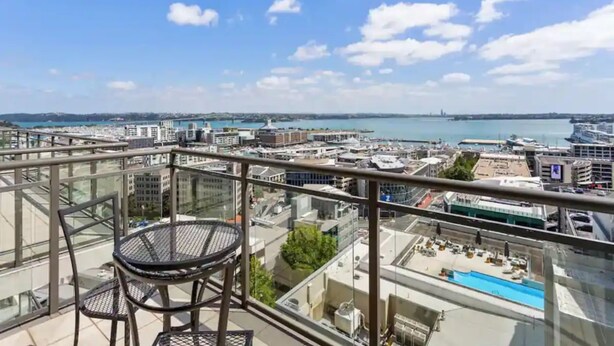 Un balcon avec une table et des chaises, donnant sur un paysage urbain avec des bâtiments et une étendue d’eau.