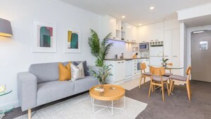 A modern apartment with a grey sofa, a round wooden coffee table, a dining area with wooden chairs, and a kitchen with white cabinets and a microwave.