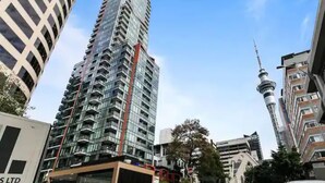 Un edificio alto con una facciata moderna in vetro, circondato da altri edifici imponenti e una torre ben visibile sullo sfondo.