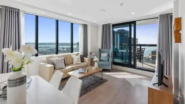 A modern living room with a sofa, coffee table, and dining table. There is a balcony with a view of the city. - Sky Tower Views | 2 Bed 2 Bath CBD | Zodiak Stays (Auckland)