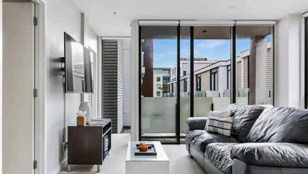 A modern living room with a grey sofa, a white coffee table, and a sliding glass door leading to a balcony. - Lovely And Cozy Apartment - Parking, Pool And Gym! (Auckland)
