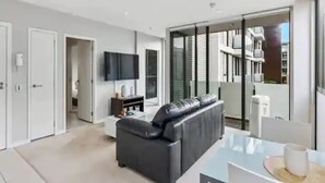 A modern living room with a black leather sofa, a flat-screen TV mounted on the wall, and a glass dining table. - Lovely And Cozy Apartment - Parking, Pool And Gym! (Auckland)